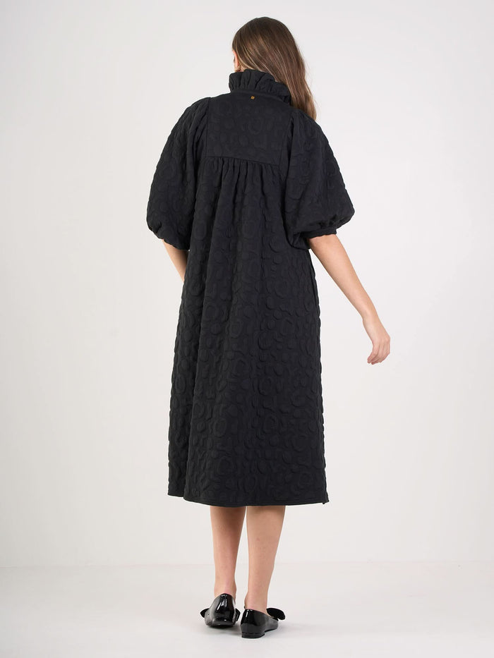 A woman stands with her back to the camera, wearing the Stella Midi dress in embossed cheetah with short puff sleeves and a ruffle stand collar, paired with black bow flat shoes, against a plain white background.