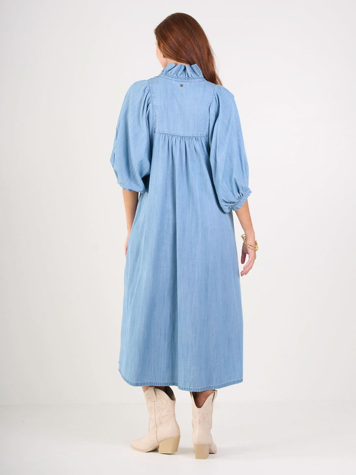 A person with long brown hair faces away, wearing the Stella Midi—a light blue denim chambray dress with a ruffle stand collar and ankle-length hem. They pair it with beige knee-high boots against a plain white background.