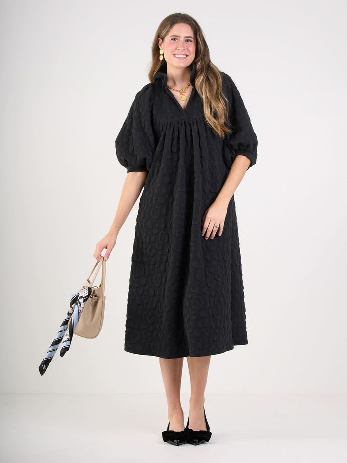 A woman wearing the Stella Midi black embossed cheetah dress with puff sleeves stands smiling, holding a beige handbag adorned with a patterned scarf. She has long hair, black shoes, and gold jewelry.