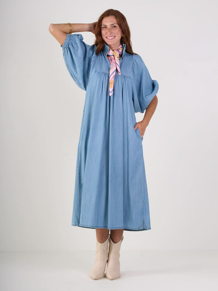 Against a plain backdrop, a person wearing the Stella Midi in blue denim chambray is paired with white boots and a colorful scarf. They are smiling, one hand on their hip and the other raised behind their head.