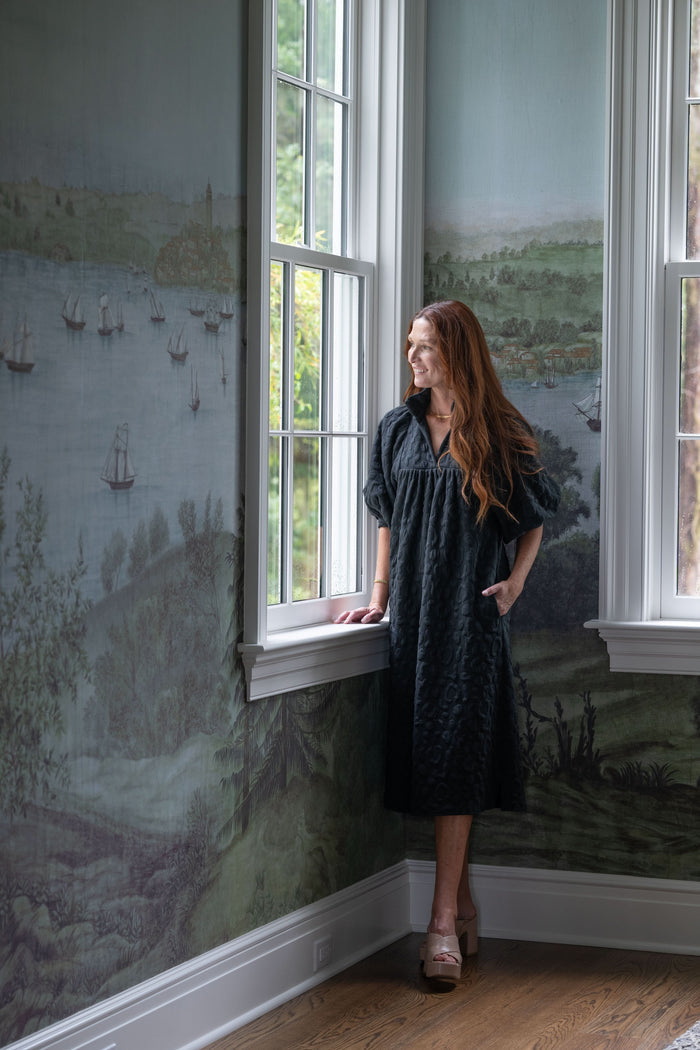 A woman in a Stella Midi - Black Cheetah dress with a ruffle stand collar stands by a large window, gazing outside. The room features walls adorned with a scenic mural depicting landscapes and boats, while the wooden floor complements the serene ambiance of the space.