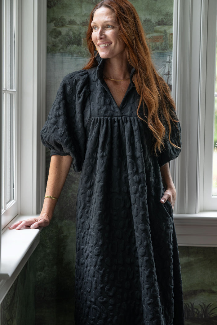 A woman with long red hair stands by a window, smiling slightly in her Stella Midi - Black Cheetah dress. She is wearing a long, textured black dress featuring short, puffy sleeves and a ruffle stand collar. The background shows a green, nature-inspired wallpaper.
