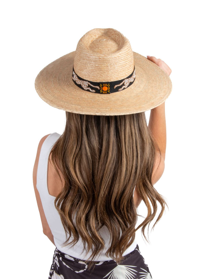 A person with long wavy hair dons the Sunshine Tienda x Emily McCarthy Chasing Cheetah Hat, which boasts a wide-brimmed straw design accented by a cheetah band. They face away, dressed in a sleeveless white top and a patterned black skirt, all set against a plain white background.
