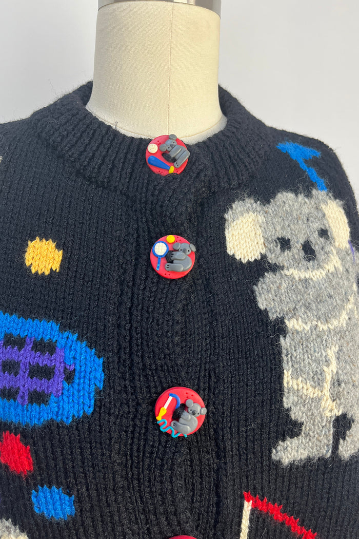 A close-up of a knitted sweater on a mannequin torso, featuring a dark navy base with a gray dog motif and colorful circular button details.