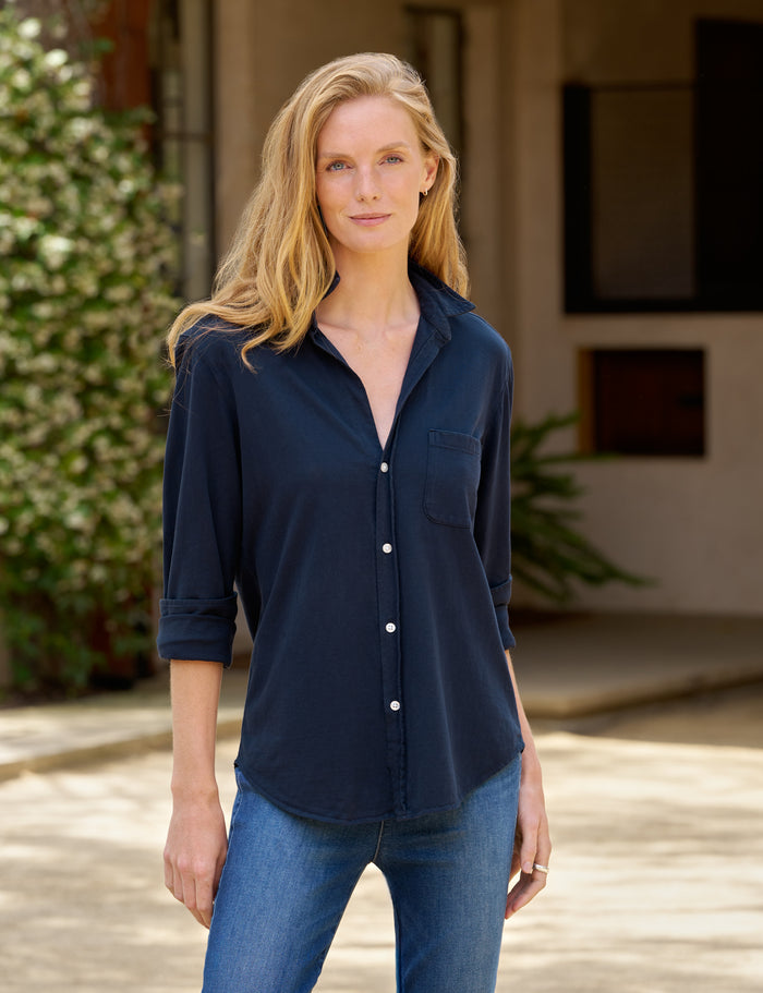 A woman wearing a dark navy blue button-down shirt with rolled sleeves and blue jeans, standing outdoors in front of a neutral-toned building.